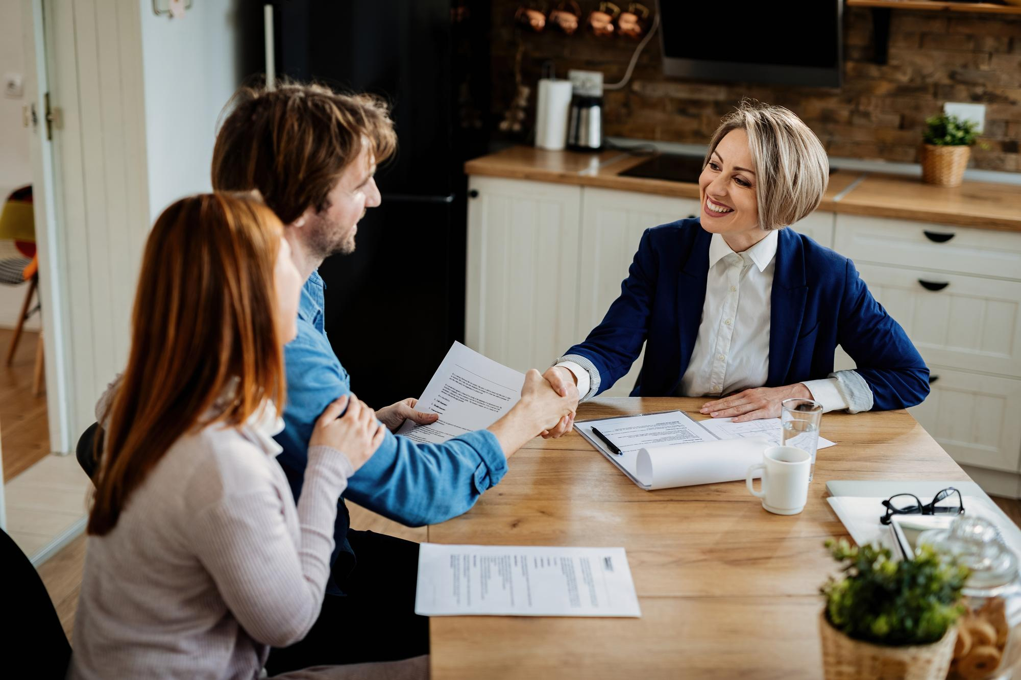 Smiling real estate agent shaking hands with a couple at a kitchen table, holding documents and offering a trustworthy, personalized home selling experience