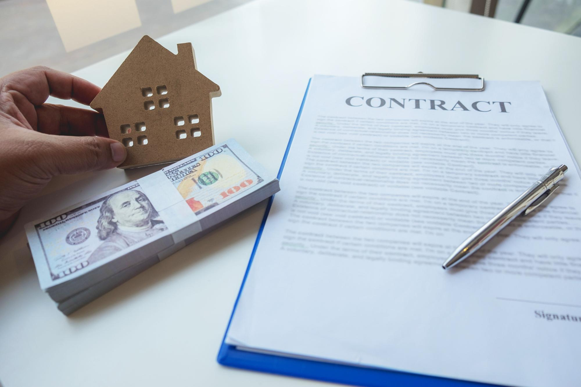 Real estate contract with house model, stack of dollar bills, and pen, symbolizing property purchase.