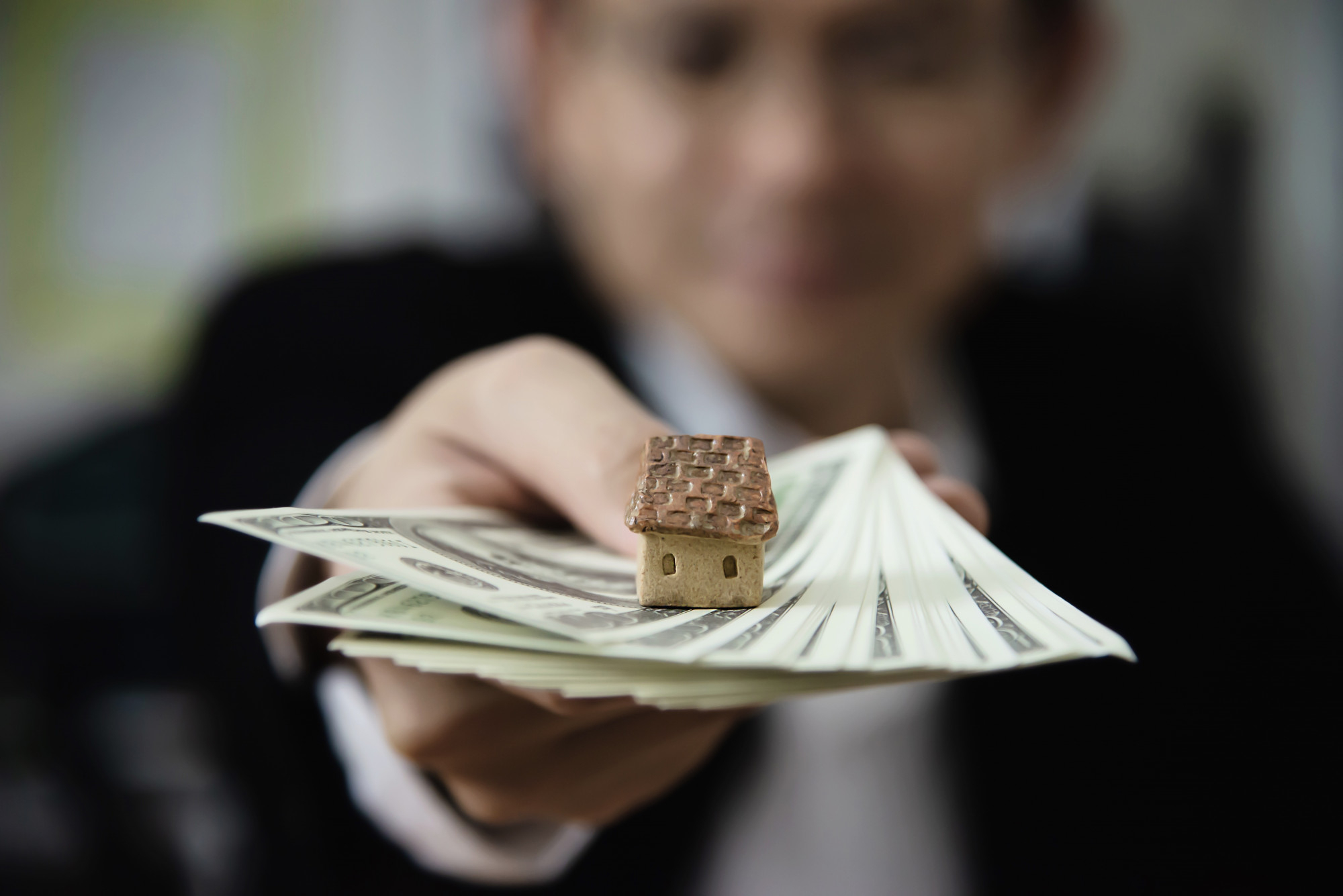 Person holding a stack of dollar bills with a small house model on top, symbolizing selling a home for cash.
