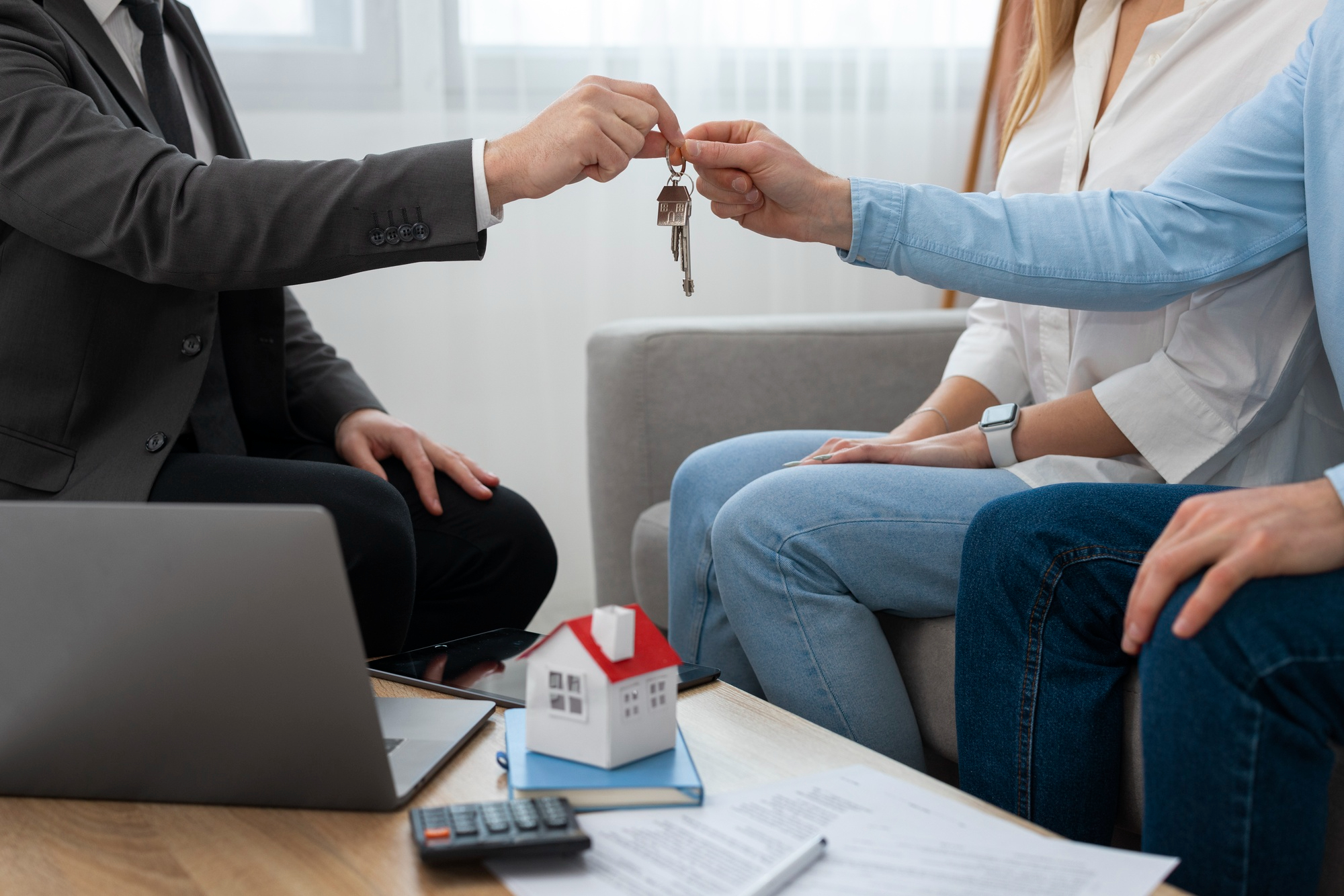 Person handing house keys to new owners, with contract, laptop, and small house model on the table, symbolizing home sale or purchase.